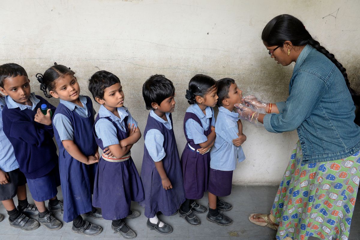An adult administers oral medication to a line of elementary school children.